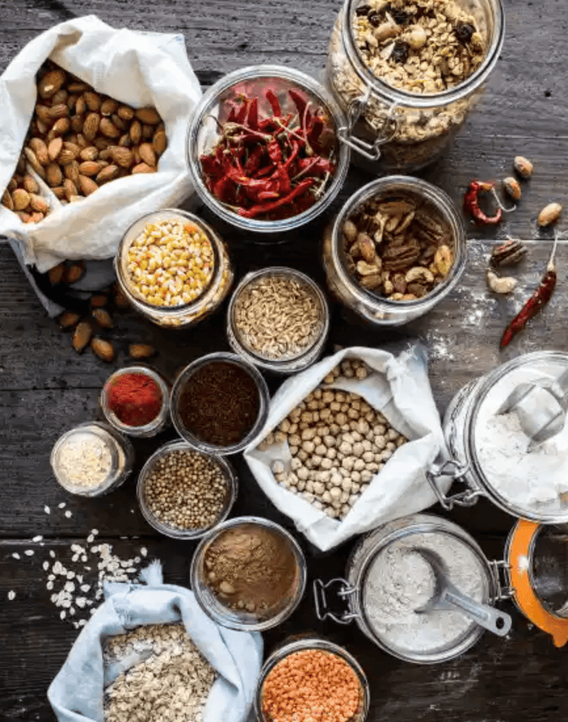 On a dark wooden table, jars and bags filled with almonds, dried chili peppers, corn kernels, chickpeas, and flour showcase Gneiss Spice’s sustainable style. Inspired by "The Zero-Waste Chef: Plant Forward Recipes and Tips for a Sustainable Kitchen," they embrace minimal plastic use.