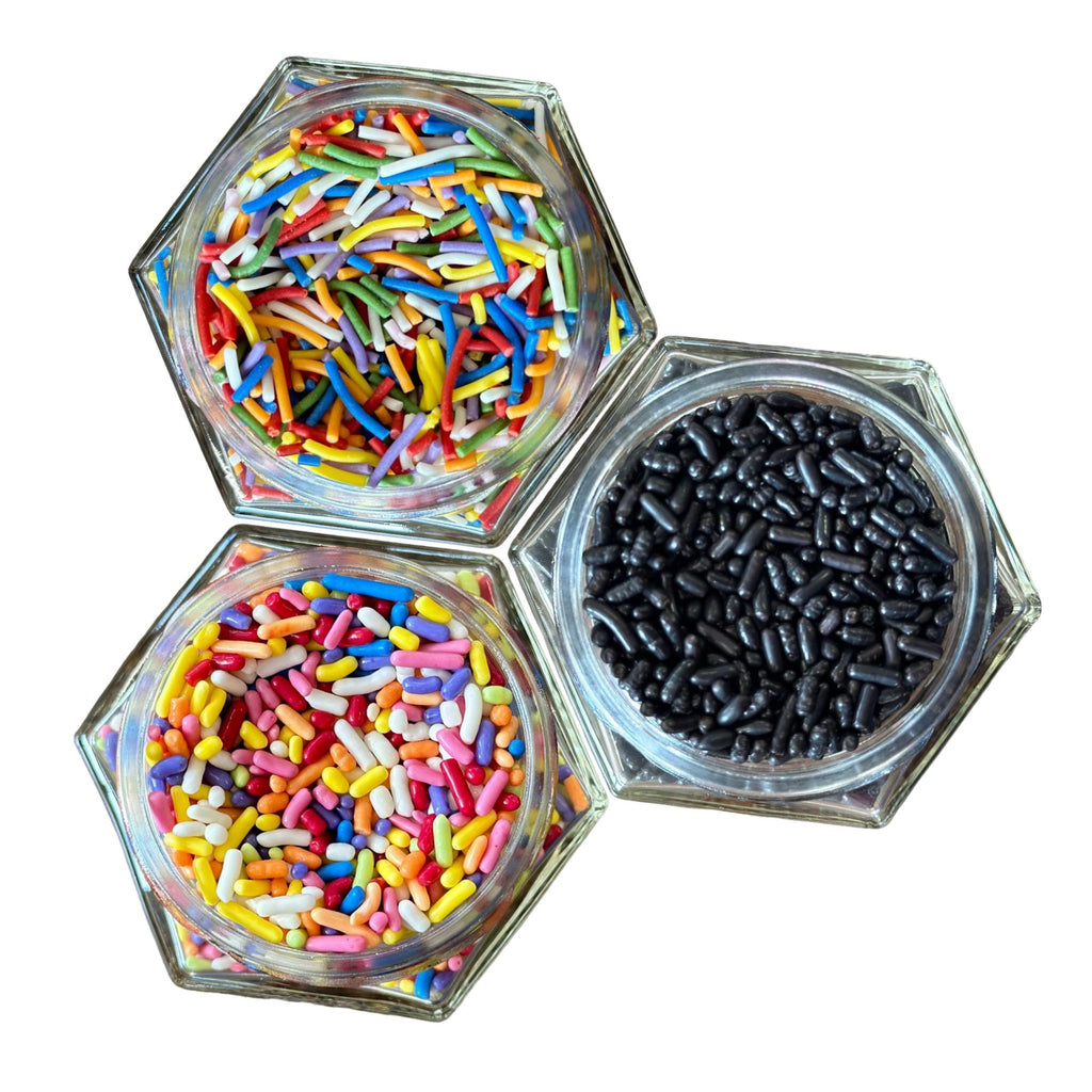 Three hexagonal glass jars from Gneiss Spice's Sprinkles 3-Pack — Dye-Free and Fair Trade Baking Decorations are arranged in a triangular pattern on a white background. Inside, you'll find vibrant sprinkles: one jar with multicolor, another in pastel hues, and the third jet black. These gluten-free sprinkles add a playful touch to any treat.