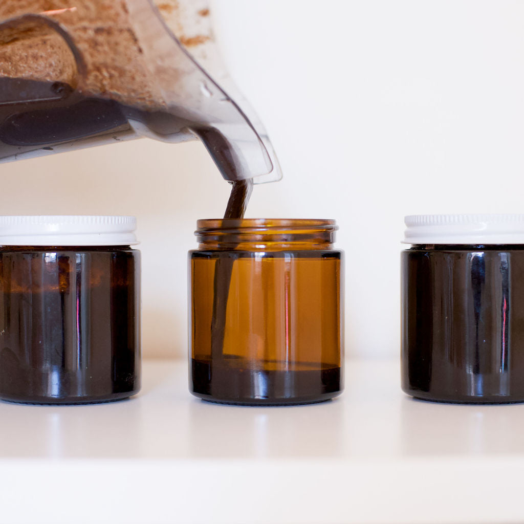 A Gneiss Spice Homemade Vanilla Paste Gift Kit blender pours a dark liquid into an amber glass jar, evoking gourmet quality. Two similar jars with white lids stand on either side, filled with the same rich essence. The plain, light-colored surface backdrop highlights the elegance of this vanilla paste masterpiece.