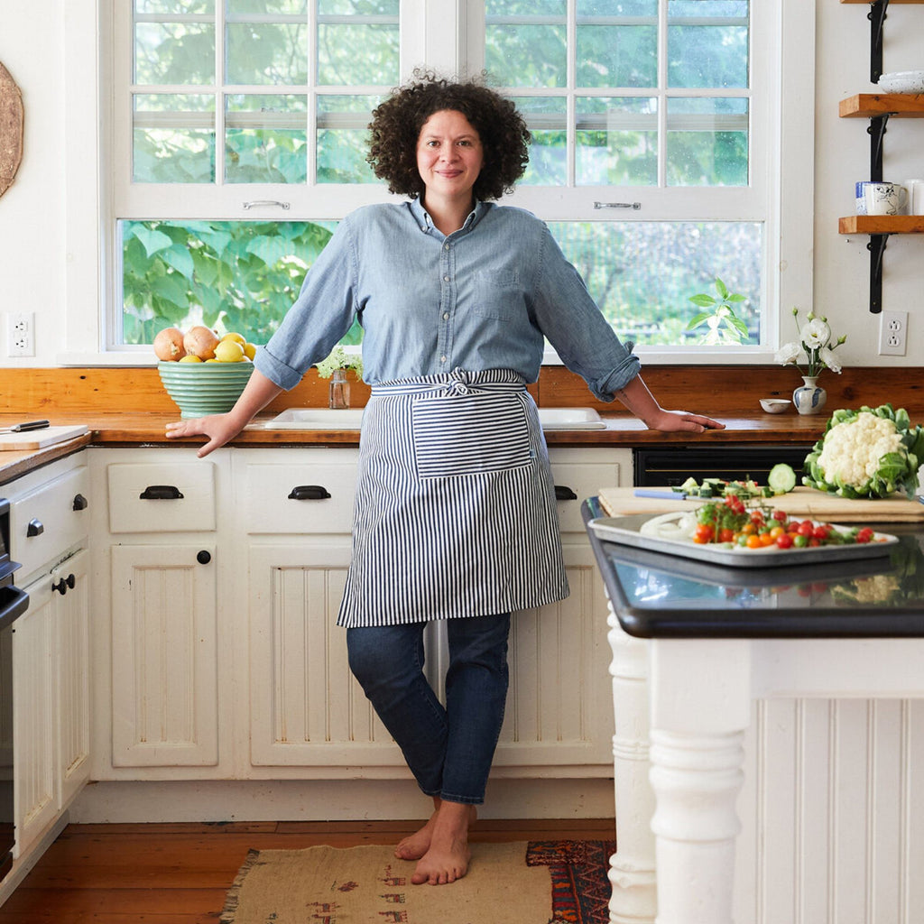 A person with curly hair stands barefoot in a bright kitchen, wearing a denim shirt and striped apron. They smile while leaning against the counter surrounded by the "What Goes With What: 100 Recipes, 20 Charts, Endless Possibilities" cookbook by Gneiss Spice on a cutting board, with assorted vegetables nearby. Large windows fill the room with natural light.