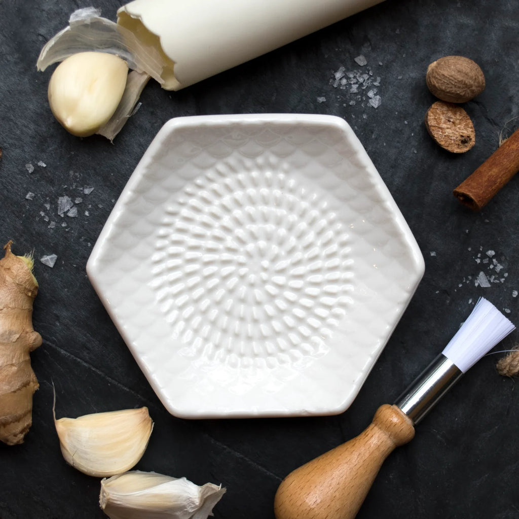 A textured white Garlic Grate Plate by Gneiss Spice sits on a dark surface, encircled by garlic cloves, ginger, a rolled parchment, nutmeg, a cinnamon stick, and a wooden-handled brush. This essential kitchen tool enhances your culinary space with both style and functionality.