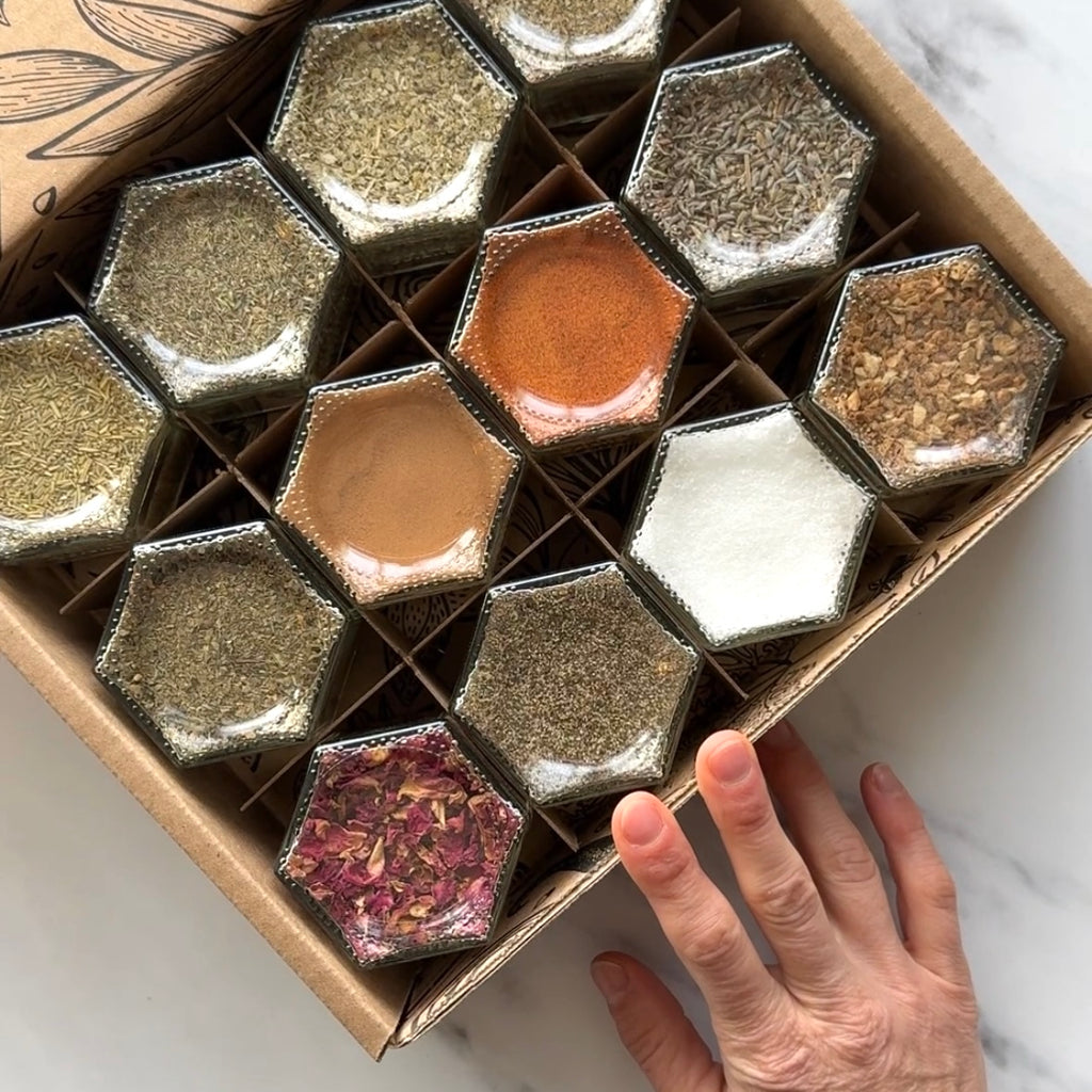 A collection of 12 magnetic hexagonal Gneiss Spice glass jars, brimming with organic spices for manifestations in vibrant red, green, and brown shades. A hand holds the edge of this New! Witches Spice Kit on a marble surface.