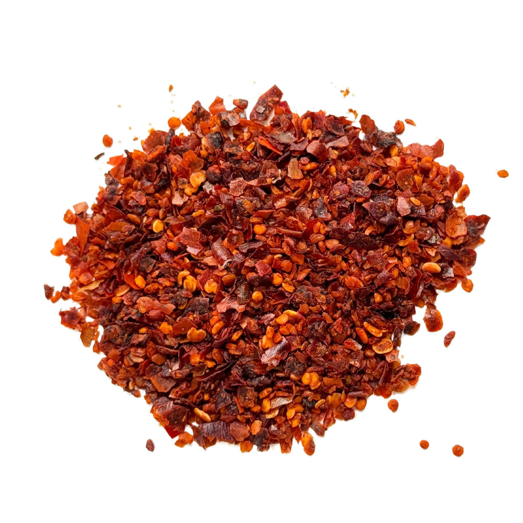 A heap of Gneiss Spice's Aleppo-Style Marash Pepper flakes on a white background, showcasing their vibrant reddish-brown color and the varied size of the flakes with visible seeds.