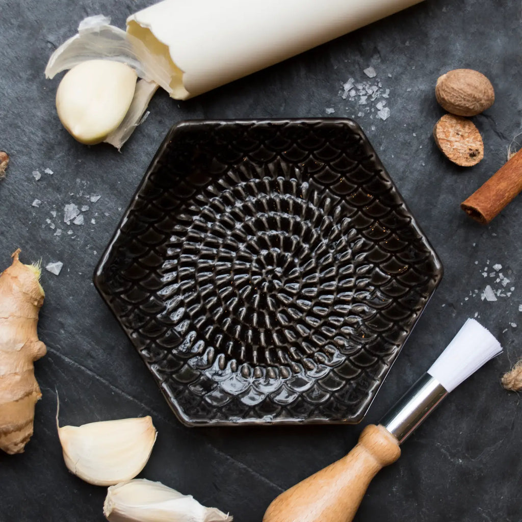 The Gneiss Spice Garlic Grate Plate, a textured black ceramic essential, is displayed on a dark surface amidst ginger, garlic cloves, whole nutmegs, a cinnamon stick, rolled parchment paper, and a wooden-handled brush—perfect for grating garlic.