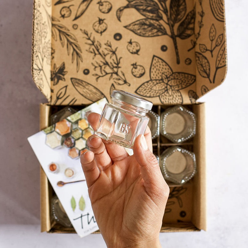 A hand holds a small, empty glass jar with a metal lid above an open box lined with botanical illustrations. Inside the Gneiss Spice 'FOODIE' Gift Bundle are magnetic jars for organic seasonings alongside a colorful hexagon-patterned gift certificate for refills.