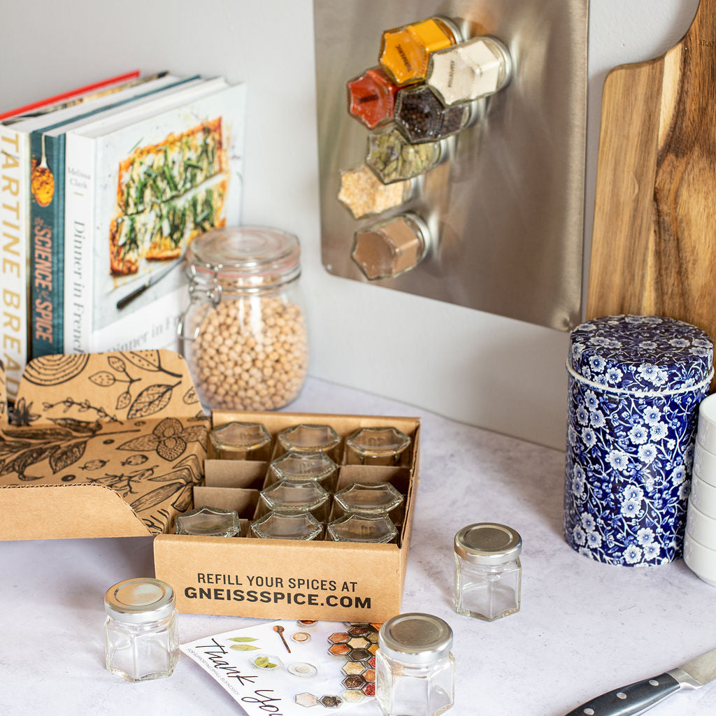 A kitchen counter showcases the 'FOODIE' GIFT BUNDLE from Gneiss Spice, featuring hexagonal magnetic jars on a board. Nearby, there are cookbooks, a jar of chickpeas, and a blue floral canister. A wooden cutting board rests next to a box inviting you to refill your organic seasonings at GneissSpice.com with a gift certificate for organic refills.