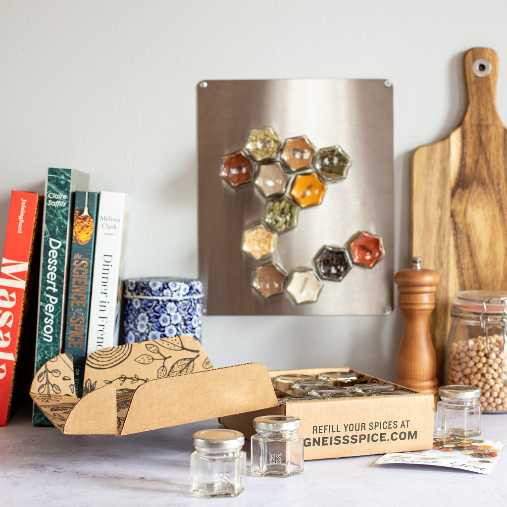 A kitchen countertop showcases a spice rack on the wall with hexagonal magnetic jars from Gneiss Spice. The counter features a wooden cutting board, a pepper grinder, and cookbooks, along with a partially open cardboard box containing the 'FOODIE' GIFT BUNDLE: Empty Magnetic Jars & Gift Certificate for Organic Refills.