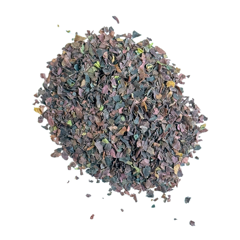 A scattering of Gneiss Spice's Dulse Flakes from the Gulf of Maine Seaweed, featuring a blend of dark purple, green, and brown hues on a white background. This nutrient-rich product provides a rich umami flavor.