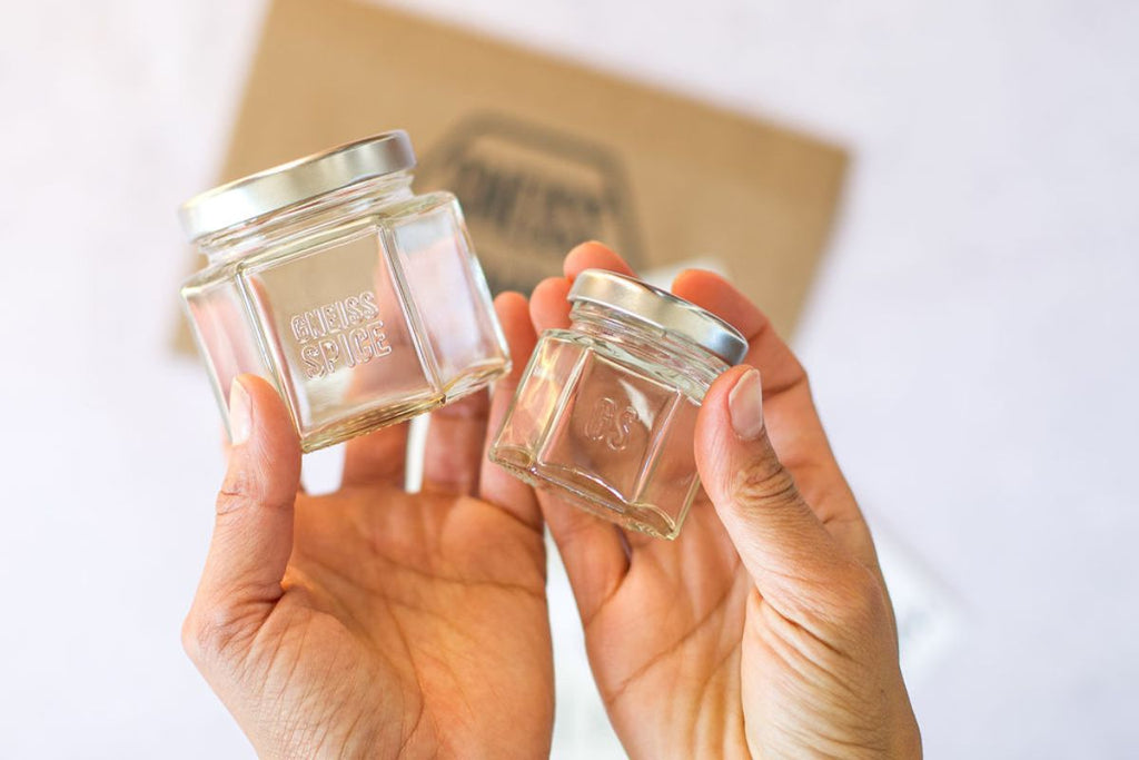 Two hands hold two small, clear glass jars with silver lids. One jar is larger with "Endless Spice" embossed on it. A blurred brown package is in the background.