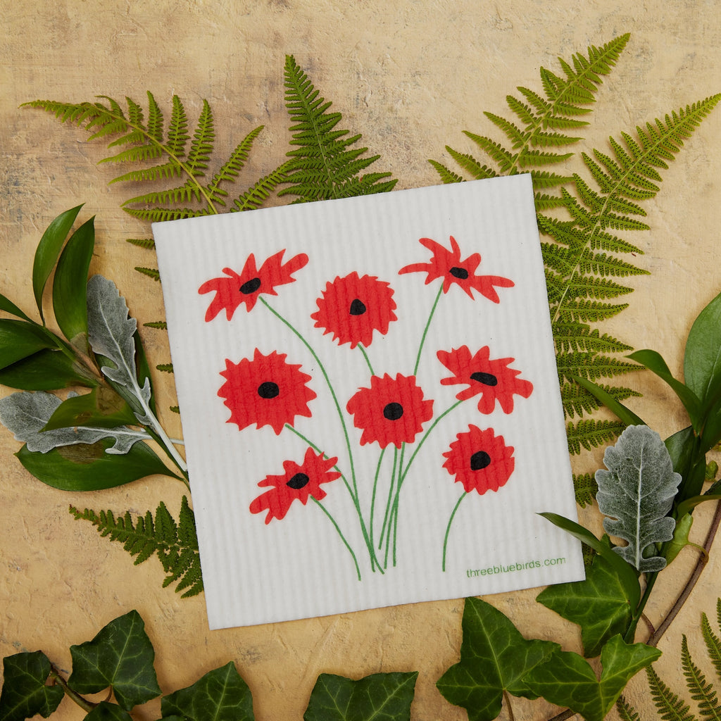 A Swedish Dishcloth from Gneiss Spice, adorned with red flowers and green stems, rests on a textured surface. This eco-friendly setup is enhanced by surrounding fern and ivy leaves, providing a sustainable option for natural decor enthusiasts. Don't forget their promotion: Buy 3, Get 1 Free!