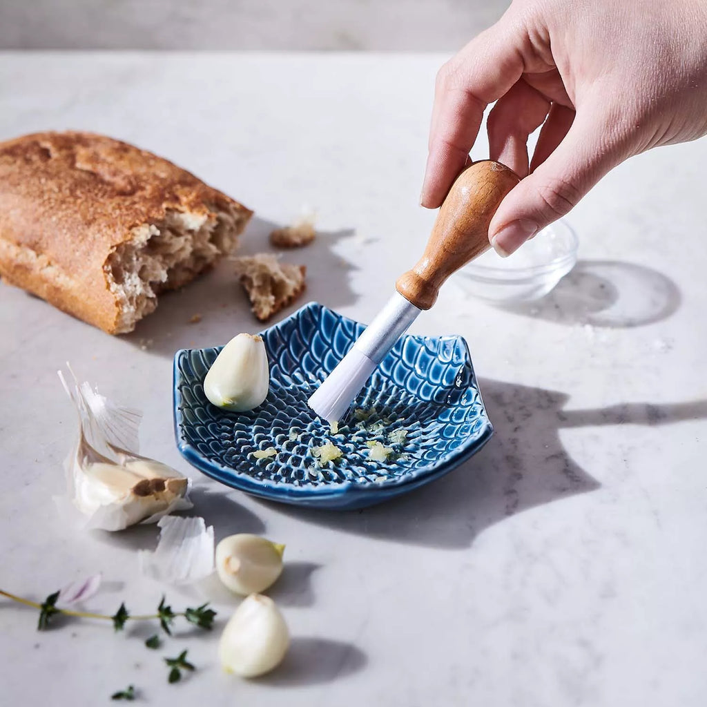 Using the Gneiss Spice Garlic Grate Plate, a hand grates garlic cloves with a wooden-handled tool on its blue ceramic surface. Nearby on the marble countertop, a partially eaten loaf of bread, garlic cloves, and sprigs of herbs are displayed. This stoneware dish is dishwasher-safe for convenient cleaning.