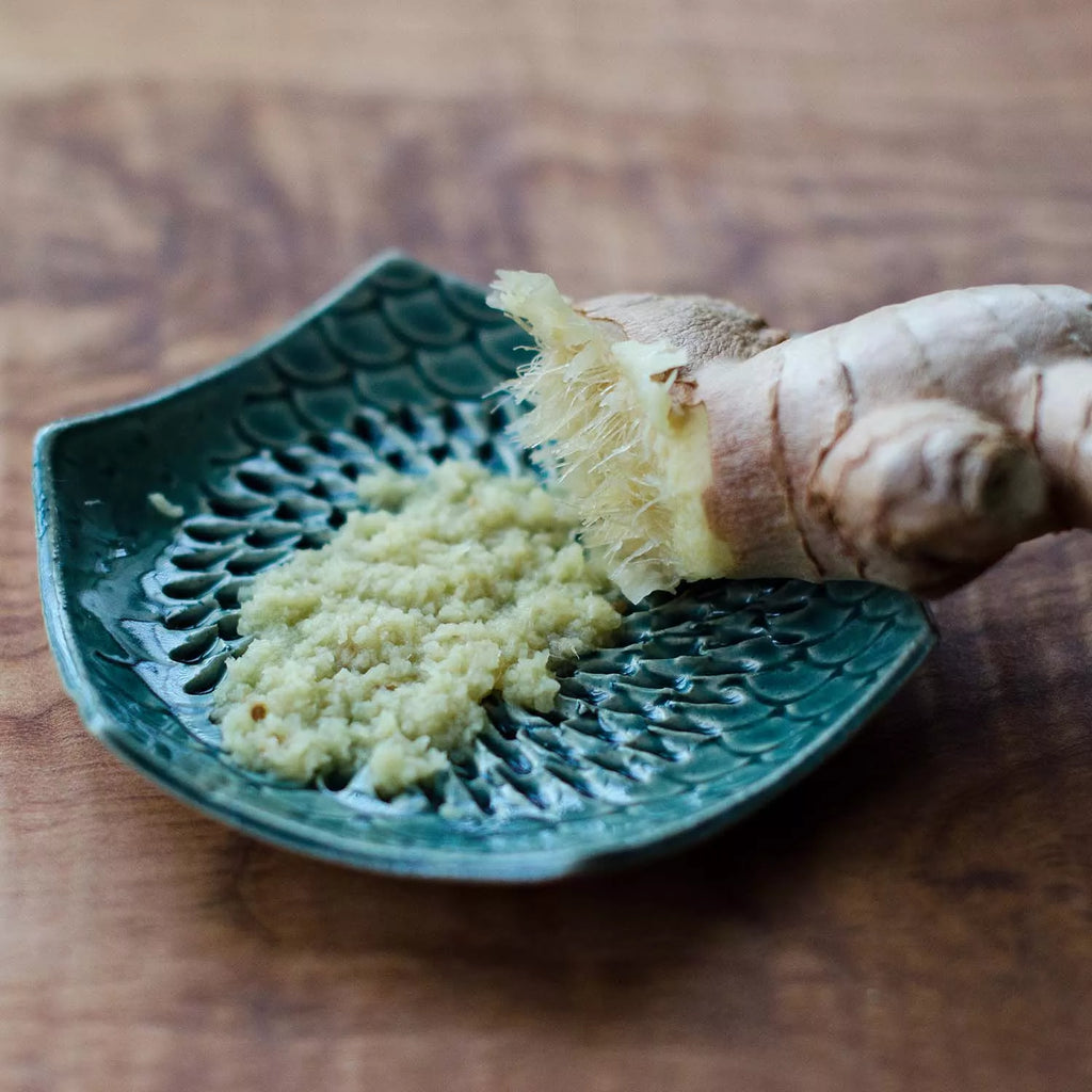 A close-up of a ginger root and freshly grated ginger on the Garlic Grate Plate from Gneiss Spice. The grating plate's intricate design enhances the contrast against the smooth wooden surface, and as part of its features, this stoneware is dishwasher-safe.