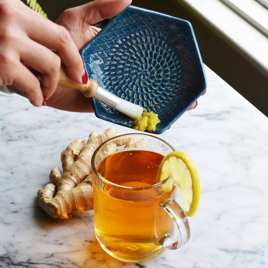 A person grates ginger onto a Garlic Grate Plate by Gneiss Spice over a marble surface. Beside the dish, a glass mug of golden tea is garnished with a lemon slice, with fresh ginger root lying next to it. Sunlight streams in from the window.