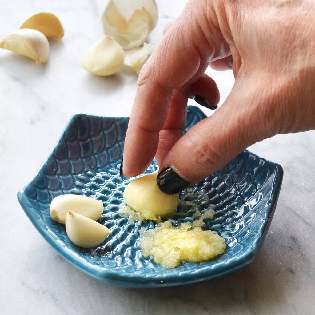 A hand skillfully grates a garlic clove on the Gneiss Spice Garlic Grate Plate, turning it into a fine paste. Several additional cloves are close by, with more pieces scattered across the marble countertop in the background. Being dishwasher-safe stoneware makes cleanup easy after your culinary creations.