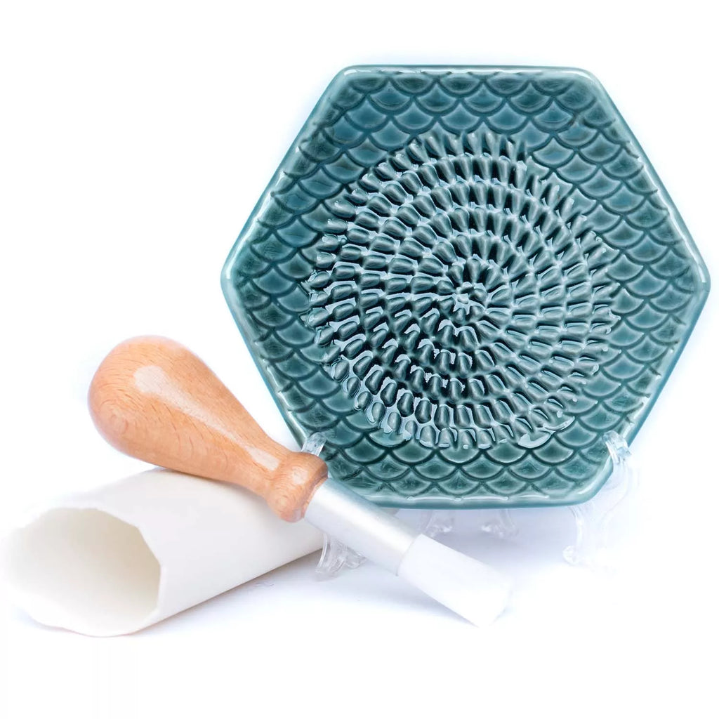 The Garlic Grate Plate by Gneiss Spice is a hexagonal ceramic tool in green, featuring a textured pattern for effective grating. It includes a pestle with a wooden handle and comes with a white silicone tube designed for peeling. Crafted from stoneware, it's dishwasher-safe for effortless cleaning.