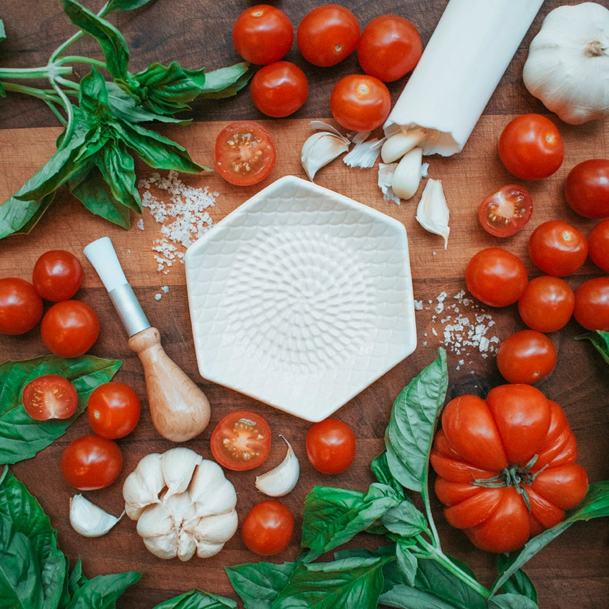 The Garlic Grate Plate by Gneiss Spice, a hexagonal dish made from dishwasher-safe white stoneware, is artistically surrounded by fresh basil leaves, cherry tomatoes, a large tomato, garlic cloves, and coarse salt on a rustic wooden surface. A sleek wooden-handled tool accompanies the scene.