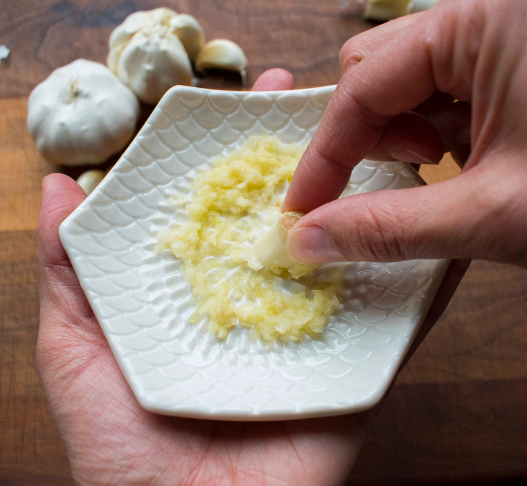 A person grates garlic on the textured surface of a Gneiss Spice Garlic Grate Plate, with whole bulbs and cloves scattered nearby on a wooden table. The finely minced garlic rests delicately on the dishwasher-safe ceramic stoneware.