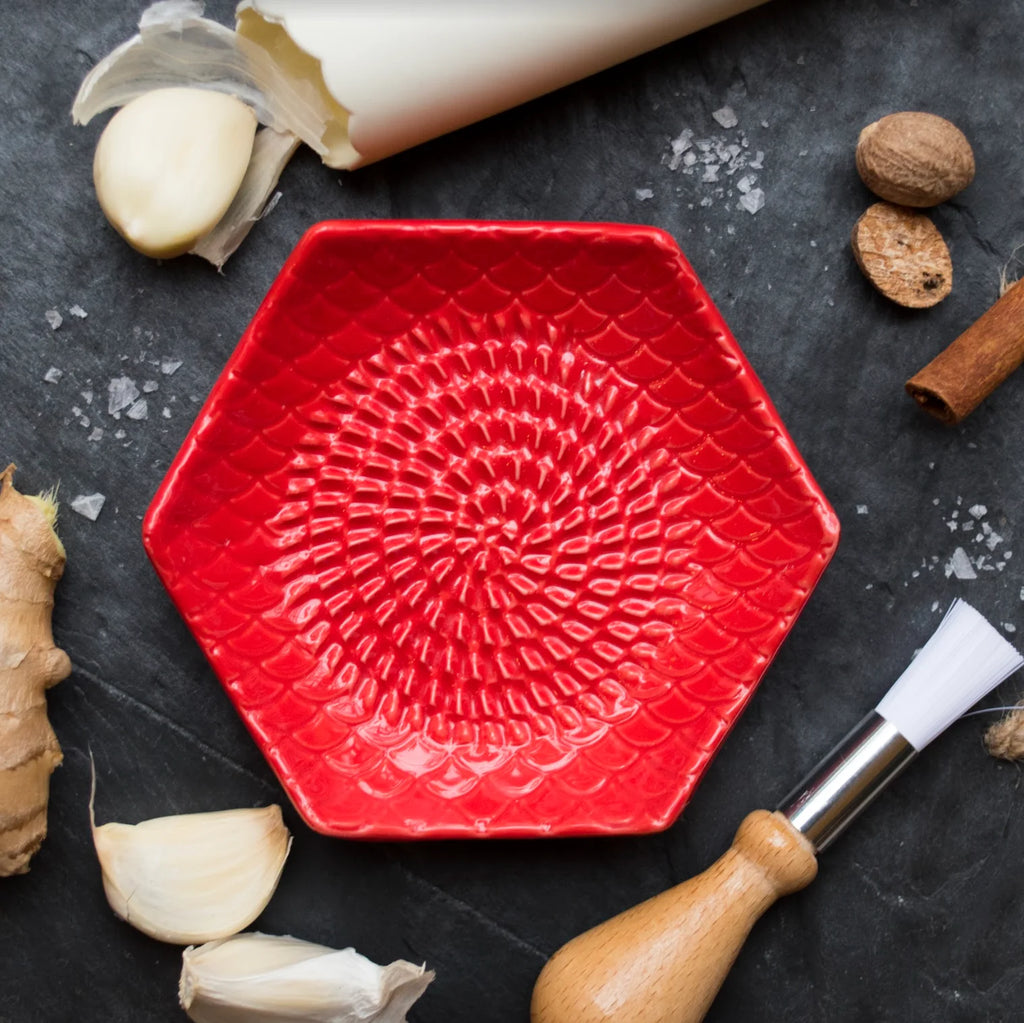 A Garlic Grate Plate from Gneiss Spice, featuring a textured pattern and hexagonal shape in red ceramic, is placed on a dark surface. Surrounding this kitchen tool are garlic cloves, ginger, nutmeg, a rolling pin, and a wooden-handled brush, with sprinkles of salt scattered around.