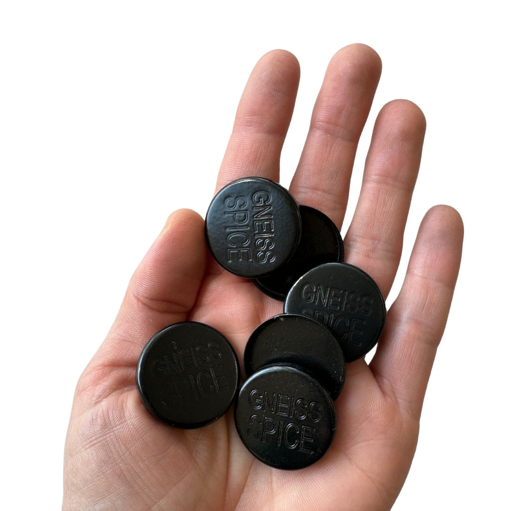 A hand holding six round, black magnet covers with the text "GNEISS SPICE" engraved on them, set against a white background. These stylish magnetic covers from Gneiss Spice are perfect for any spice jar setup.