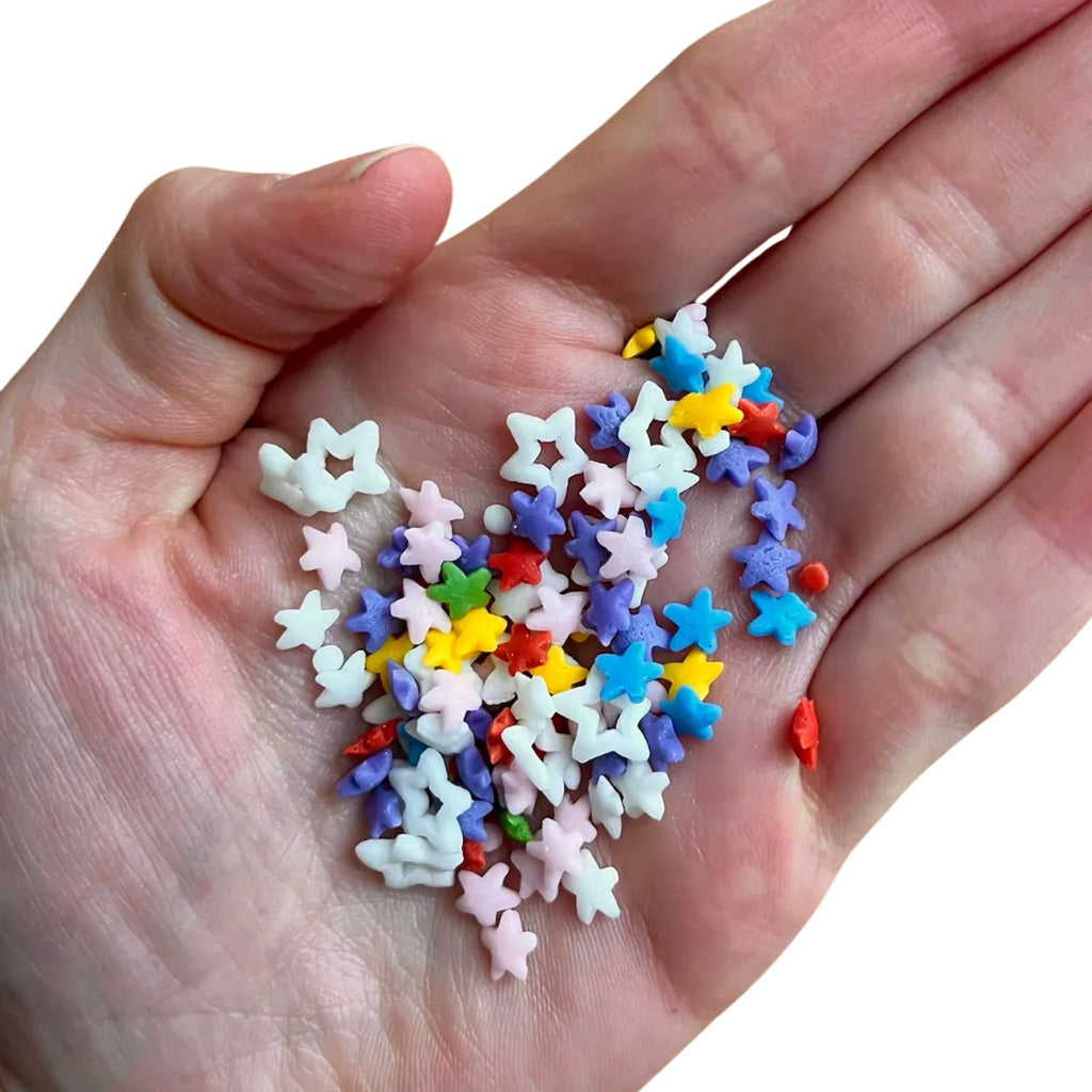 A hand cradles a vibrant assortment of Gneiss Spice's all-natural Starfetti sprinkles, featuring star shapes in pink, blue, white, yellow, and red hues. These colorful vegan sprinkles are scattered across the palm against a pristine white background.