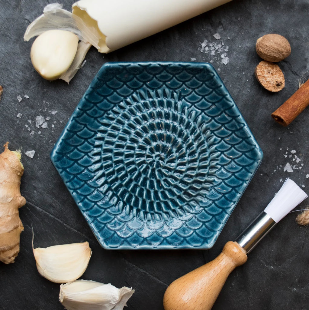 The Garlic Grate Plate by Gneiss Spice, hexagonal and textured in blue, is displayed on a dark slate surface with garlic cloves, ginger, spices, and a wooden-handled brush. This essential kitchen tool also features a roll of parchment paper that is partially visible at the top.